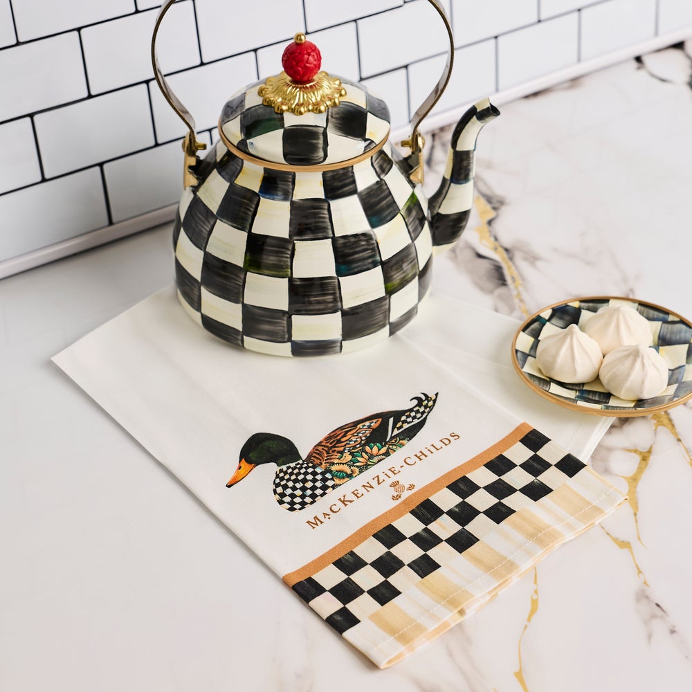 A MacKenzie-Childs checkered teapot with a gold finial sits on a white kitchen towel printed with a duck and the brand name, alongside a small dish of meringues on a marble countertop.