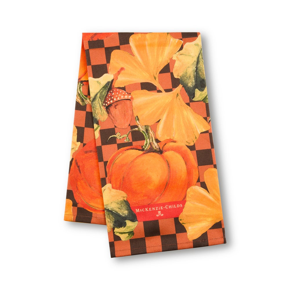 A rectangular kitchen towel featuring a fall harvest design with orange pumpkins, yellow-orange gourds, green leaves, and an acorn on an orange and black checkered background. The words 'MICKENING-CHILD'S' are printed in red at the bottom.
