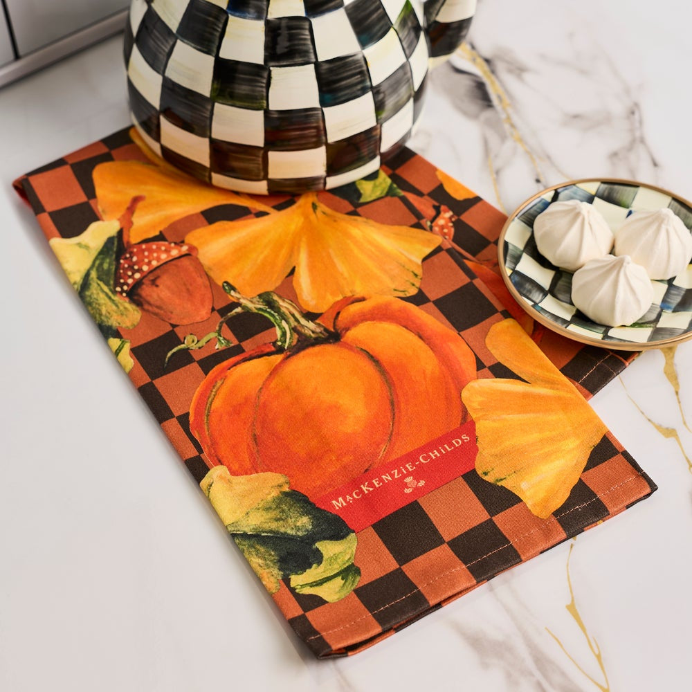 A rectangular table runner with a pumpkin and acorn autumn print, styled on a white marble surface with a black-and-white checkered teapot and a small bowl of white meringue.