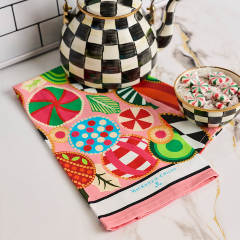 A folded pink and multicolor patterned cotton tea towel with the brand name 'MCKINNEY & LEE' printed on the border, styled on a marble surface next to a black and white checkered teapot and a bowl of candy-coated pretzels.