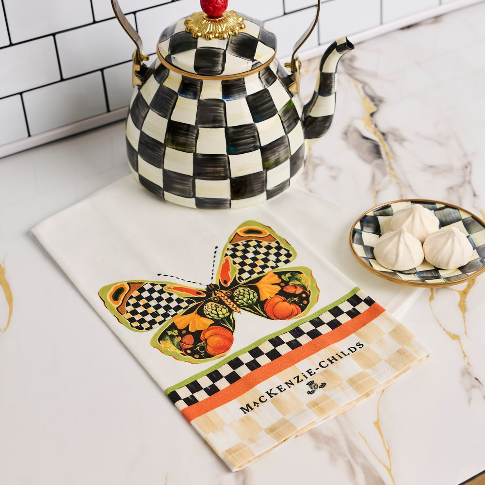 A decorative tea towel with a butterfly print and the words 'MADE IN CHILE' laid on a white marble surface, styled with a black-and-white checkered teapot and a small dish of sugar.
