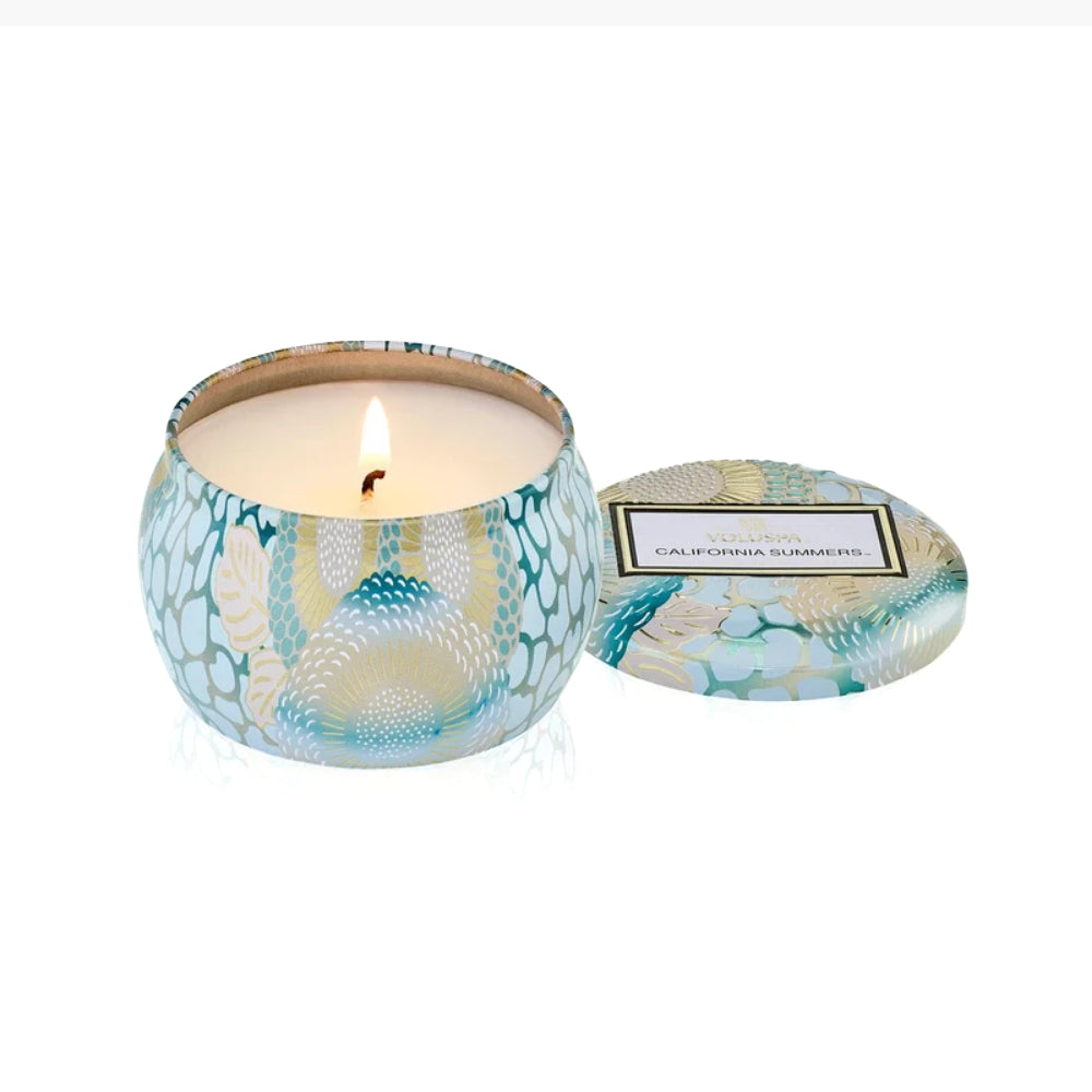 A lit scented candle in a round patterned tin with its matching lid placed beside it on a white background. The tin features a teal, blue, and cream floral and leaf design, and the label reads 'Scent of Calypso' and 'California, Sumatra'.