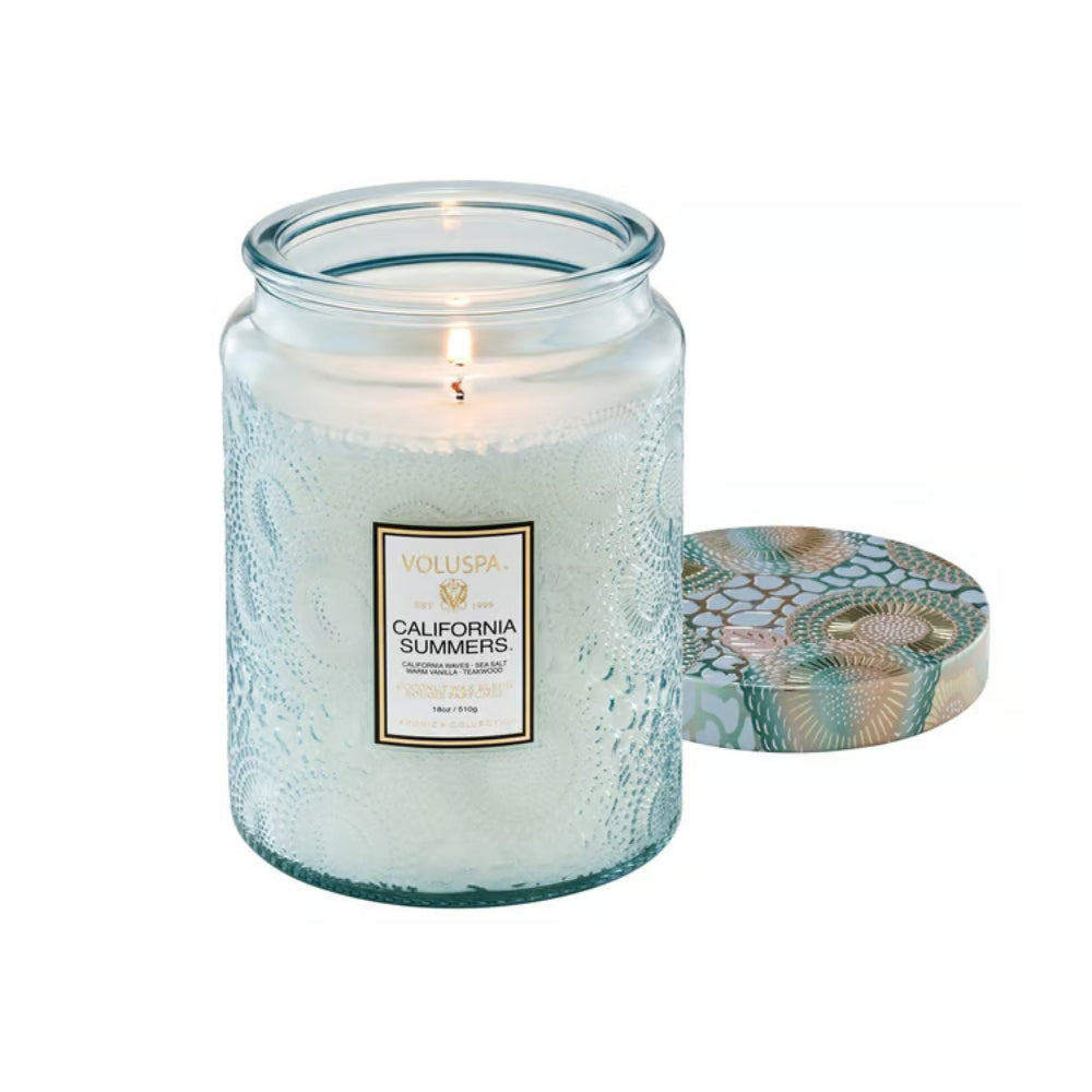 A lit Voluspa California Summers scented candle in a textured clear glass jar, shown next to its lid which features a blue and gold patterned design, on a solid white background.