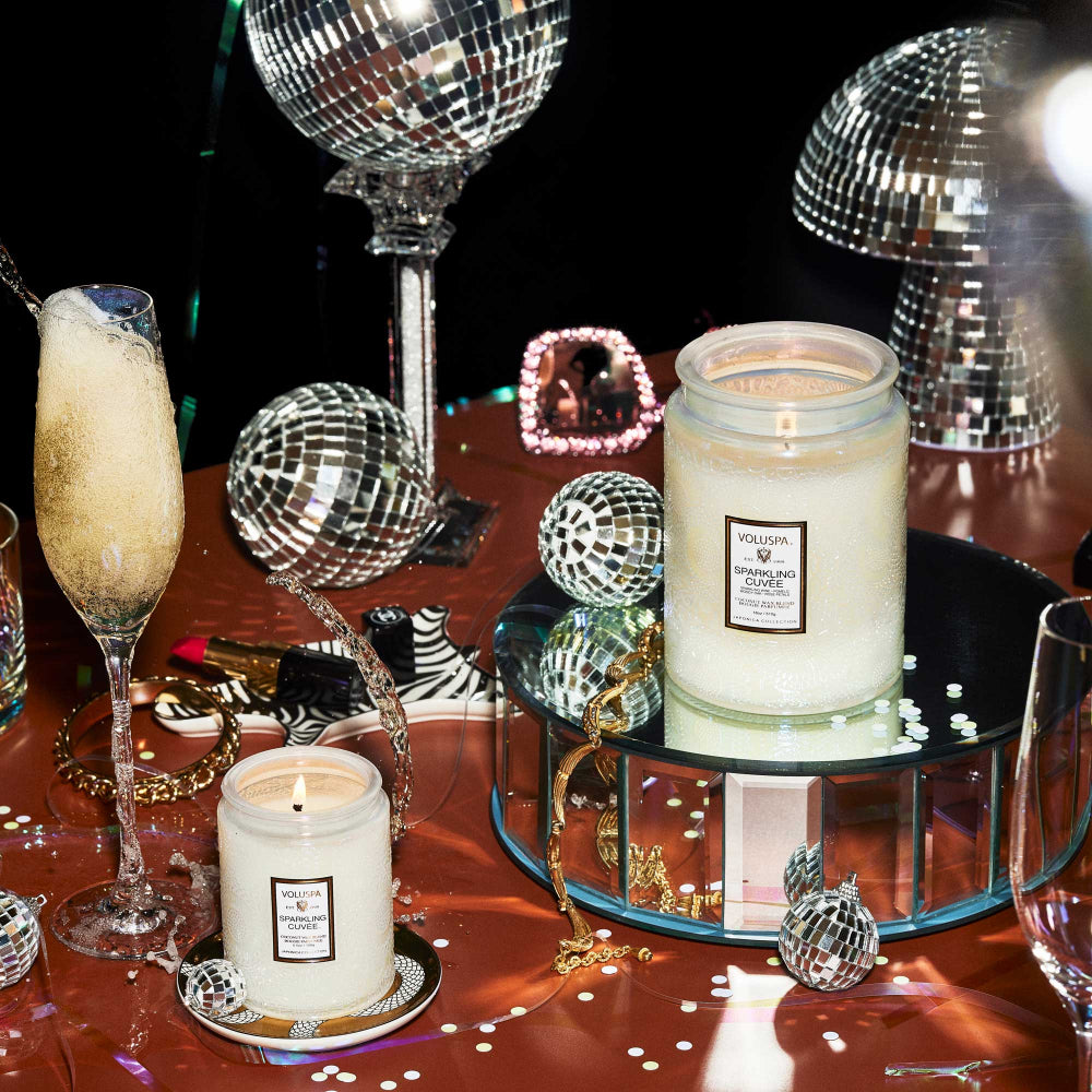 Small white Sparkling Cuvee candle on a brown table with disco balls and wine glasses. 