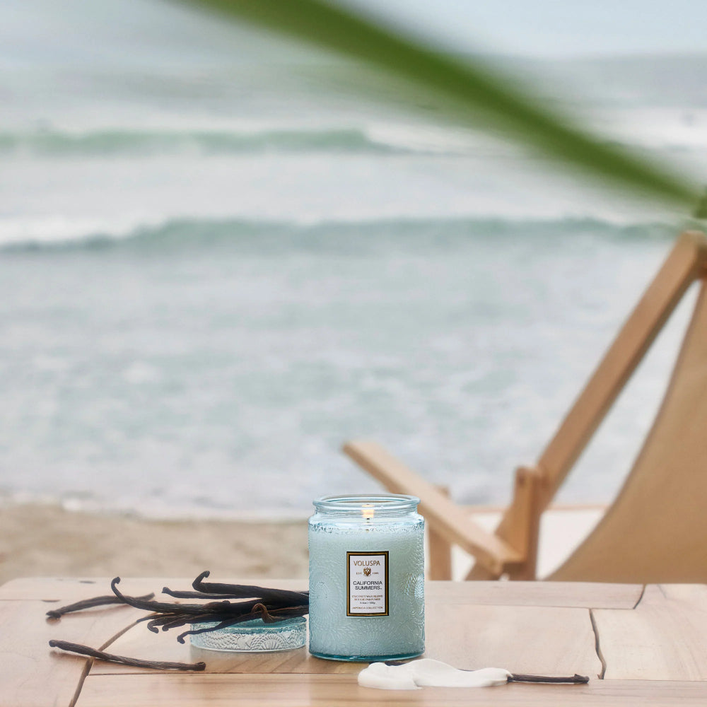 A lit candle in a clear glass jar sits on a wooden table at the beach, with vanilla beans, a small dish, and a white flower in the foreground. The ocean and a wooden chair are blurred in the background.
