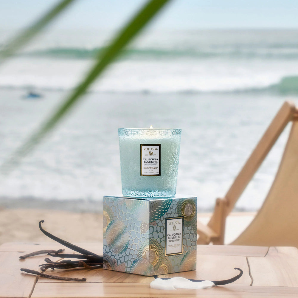 A light blue candle in a textured glass jar sits on top of a matching decorative box on a wooden table, with a blurred beach and ocean background and vanilla beans nearby.