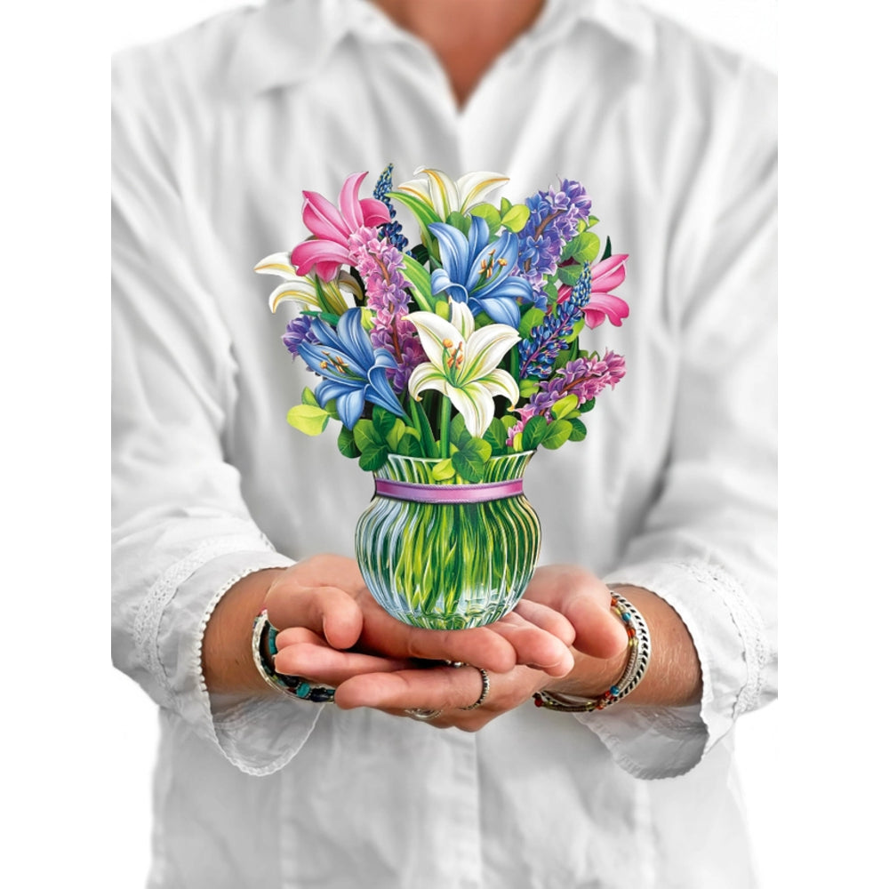 A person in a white shirt holds a vibrant, colorful bouquet of artificial flowers in a green and blue striped glass vase with a pink ribbon, set against a plain white background.