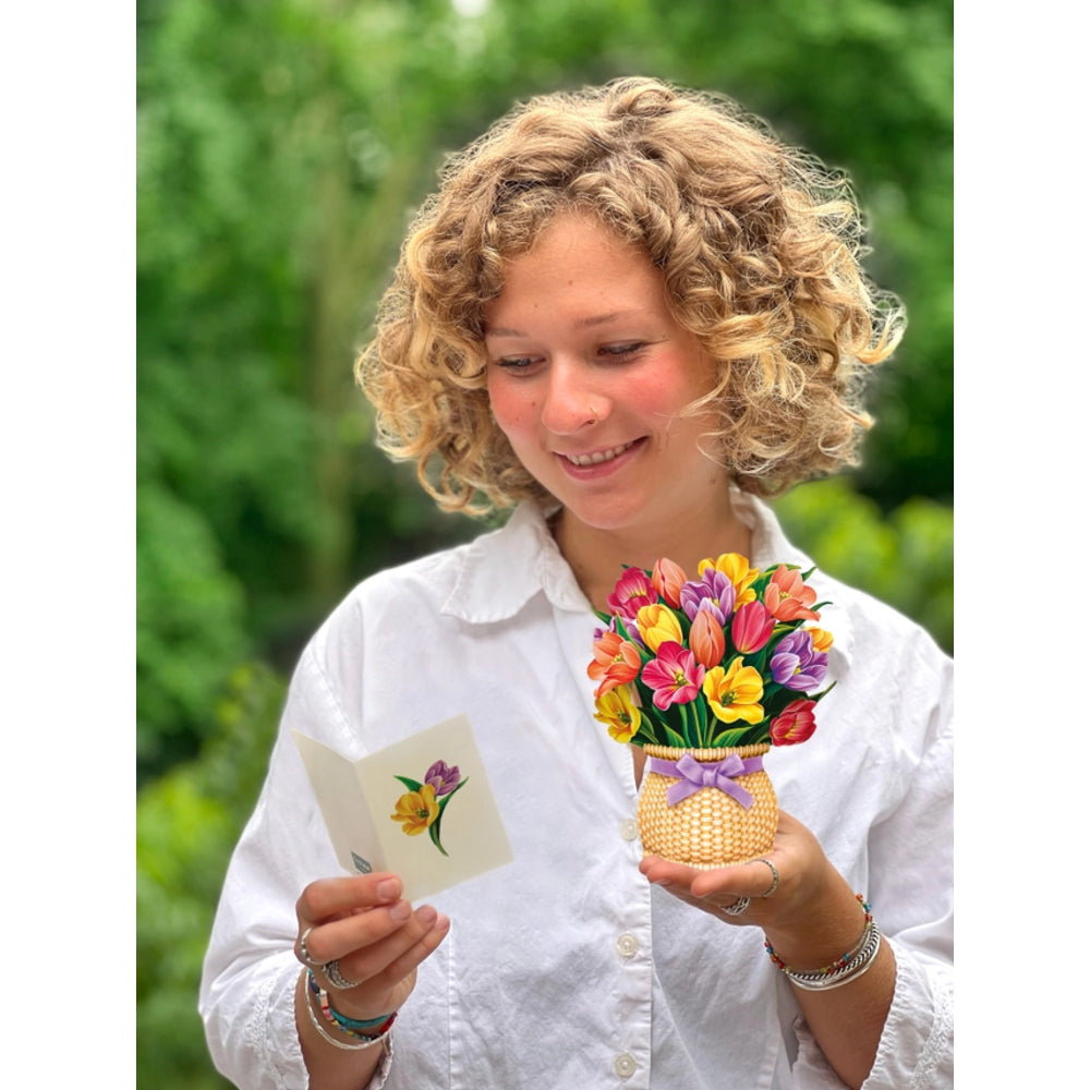A smiling woman with curly blonde hair, wearing a white button-up shirt, holds a small card with a yellow and purple flower illustration in one hand and a small woven basket vase filled with colorful artificial tulips in the other, set against a soft-focus green outdoor background.