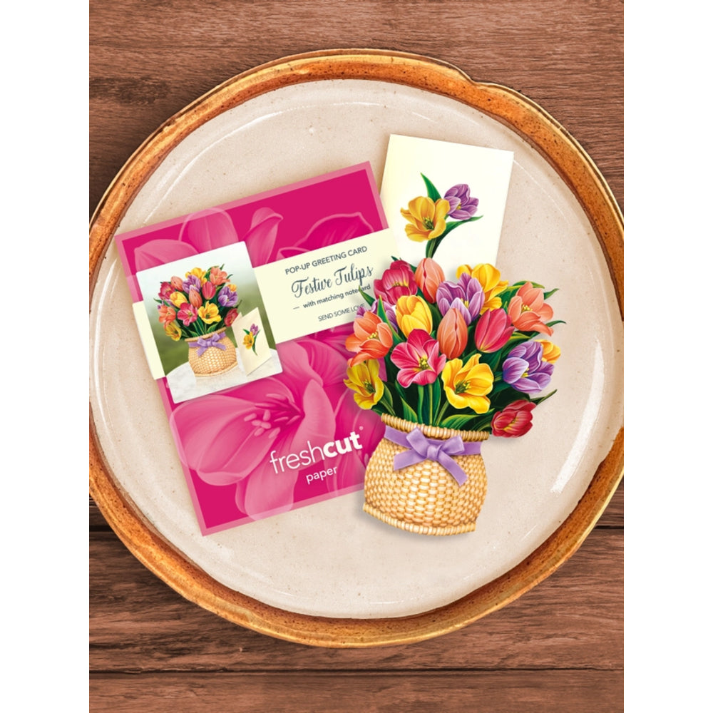 A flat-lay product shot of a 'Fallen Tulips' pop-up greeting card set by Fresh Cut Paper, featuring a bright pink envelope, a printed card, and a 3D pop-up bouquet of colorful tulips, all arranged on a round ceramic plate over a wooden background.