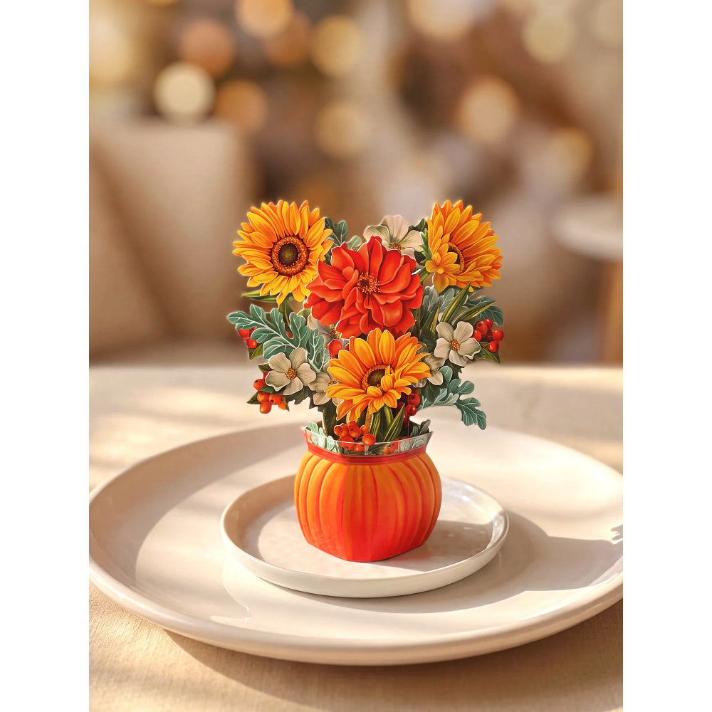 A decorative floral arrangement in a small orange pumpkin-shaped vase, placed on a white plate over a larger white platter. The bouquet includes orange and yellow flowers with green leaves and small red berries, set against a softly blurred warm indoor background.