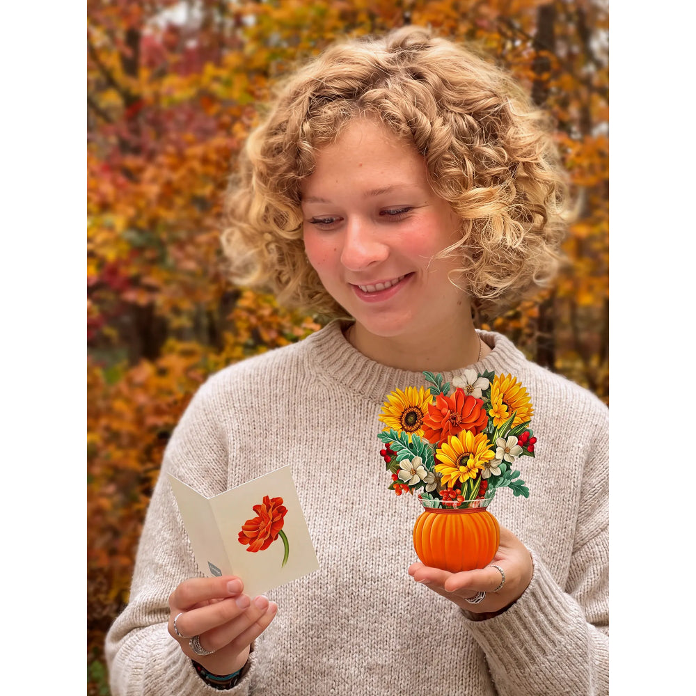A smiling woman with curly blonde hair holds a small orange pumpkin-shaped vase filled with colorful artificial flowers and looks down at a greeting card in her other hand, set against a blurred autumn garden background.