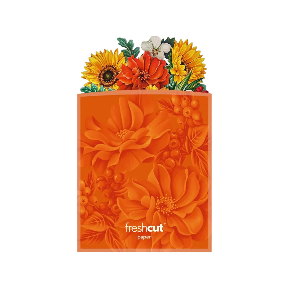 An orange Freshcut paper bag with a floral pattern, topped with a colorful bouquet of sunflowers, orange flowers, and green leaves, shown on a light gray background.