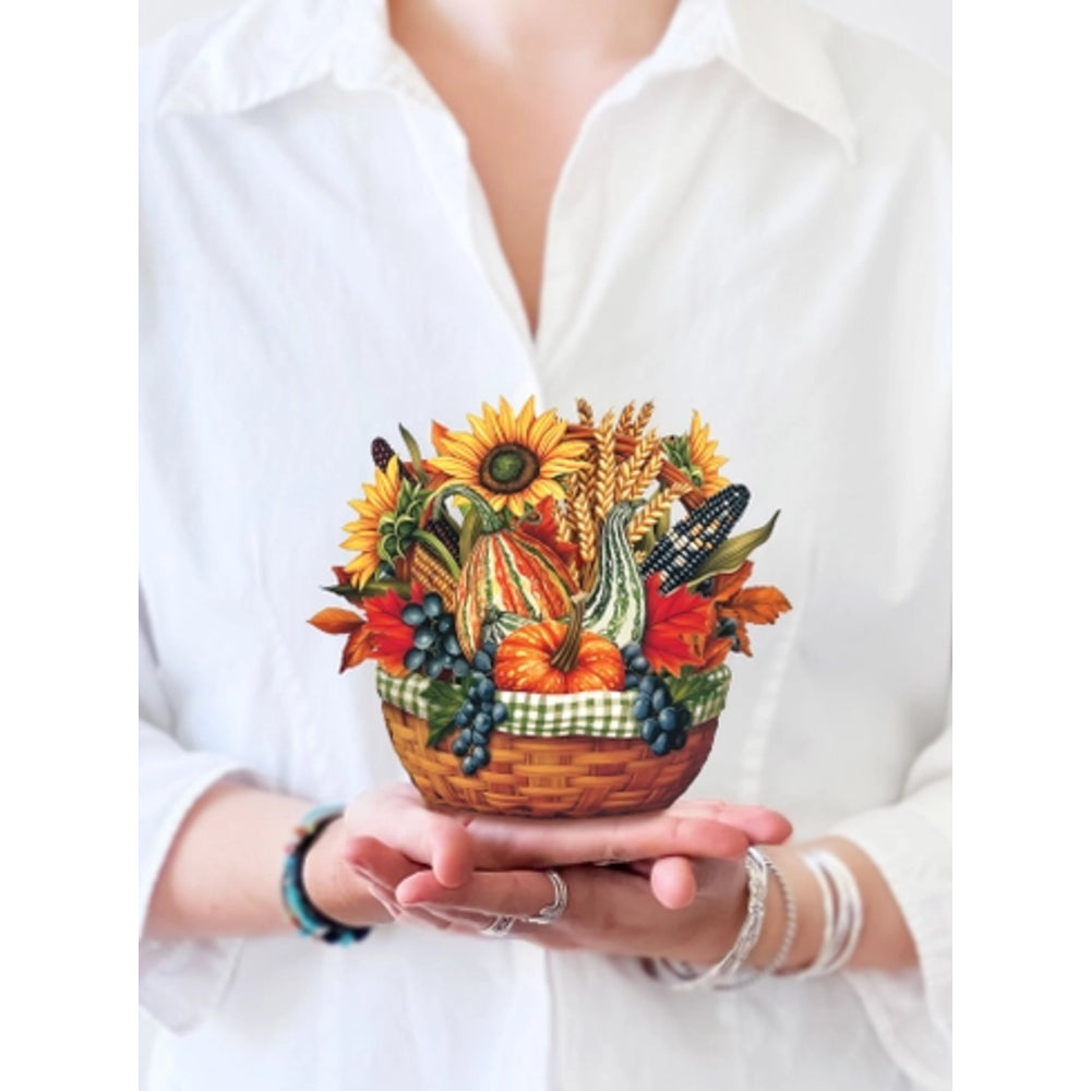 A person in a white button-up shirt holds a small illustrated basket of autumn harvest elements, including sunflowers, pumpkins, wheat, corn, leaves, and grapes, against a plain white background.