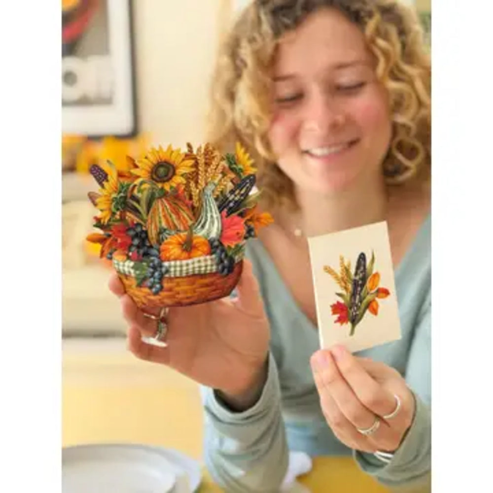 A smiling woman holds a small decorative basket filled with autumn-themed pressed flowers and a matching illustrated card in a warm indoor setting.