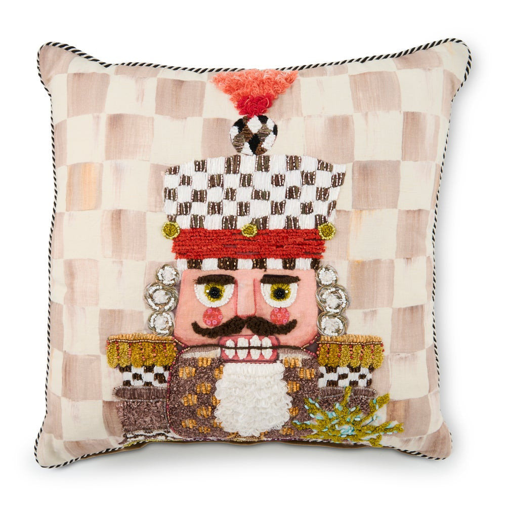 A square decorative throw pillow featuring a nutcracker character design on a beige and cream checkered fabric background. The nutcracker has embroidered facial features, a red hat with a tassel, and ornate gold and brown details, set against a clean white background.