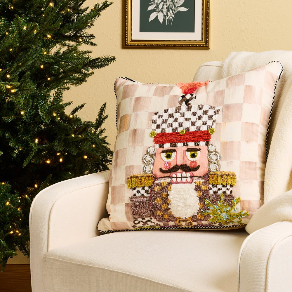 A decorative throw pillow featuring a nutcracker design, styled on a cream armchair next to a lit Christmas tree in a cozy room setting.
