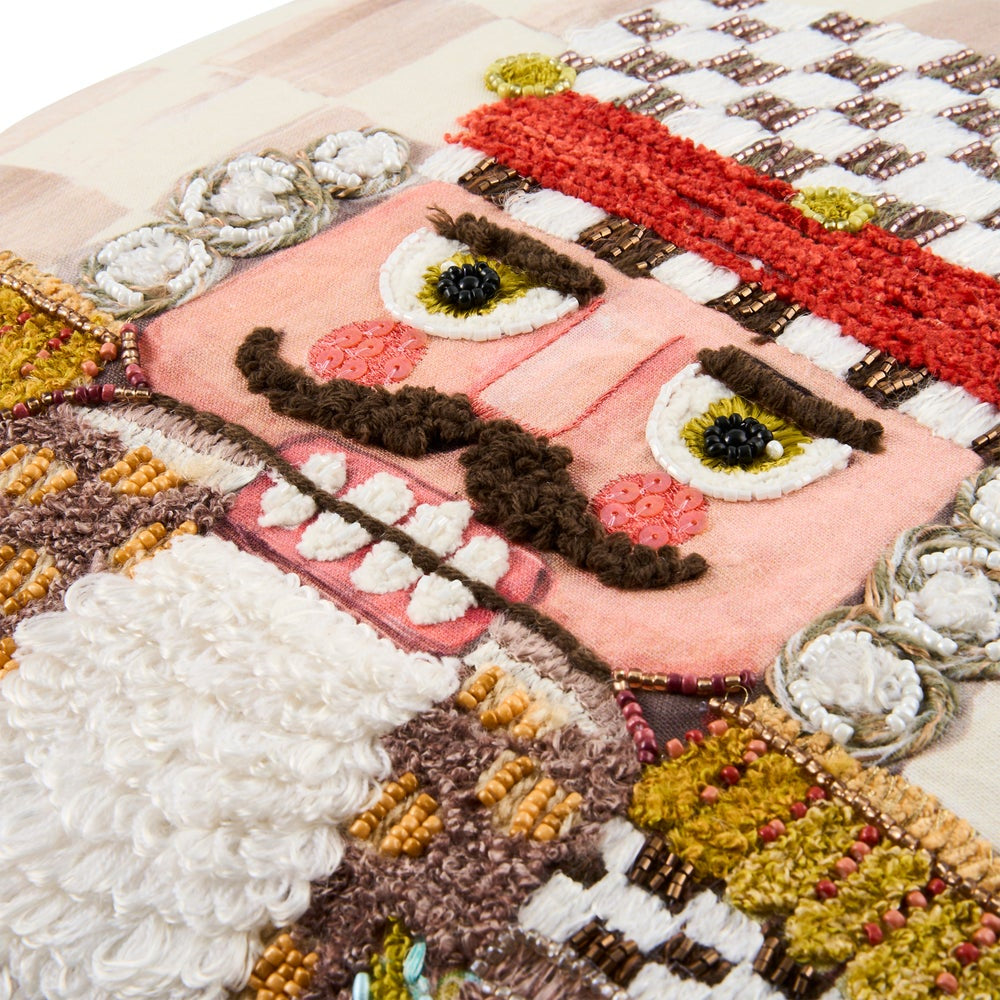 A close-up of a colorful, textured needlepoint or embroidered patch featuring a stylized face with large white and yellow eyes, a dark brown mustache, and a pink mouth, surrounded by various multicolored stitched details.