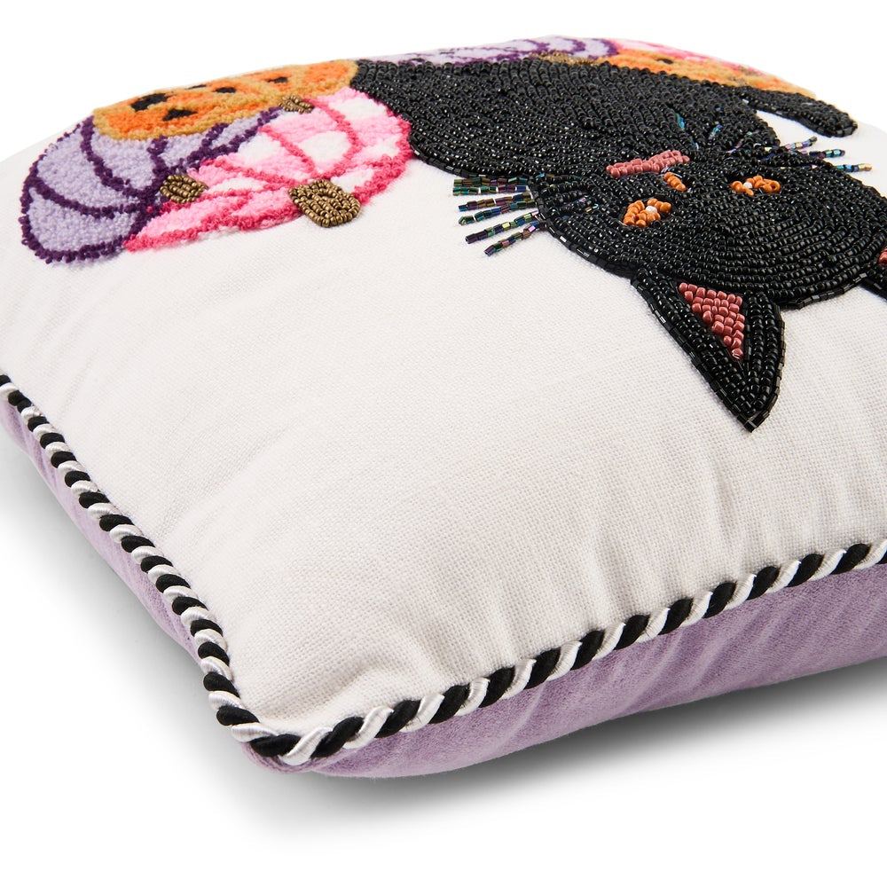 A square decorative throw pillow with a white front panel featuring a large black beaded cat and colorful floral motifs, finished with a lavender purple border and black-and-white braided piping.