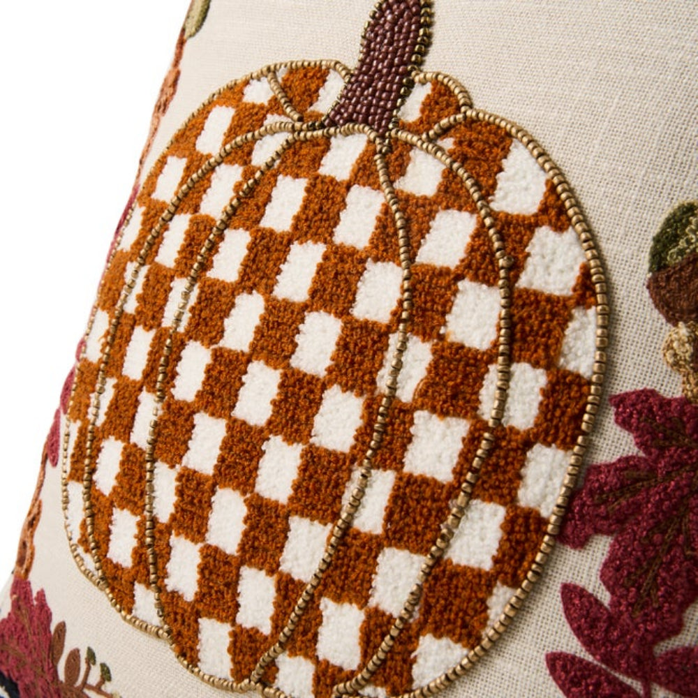 Close-up of a decorative throw pillow featuring a large autumn pumpkin motif with a brown and cream checkered pattern, gold beaded outlines, and burgundy leaf accents on a light beige fabric background.