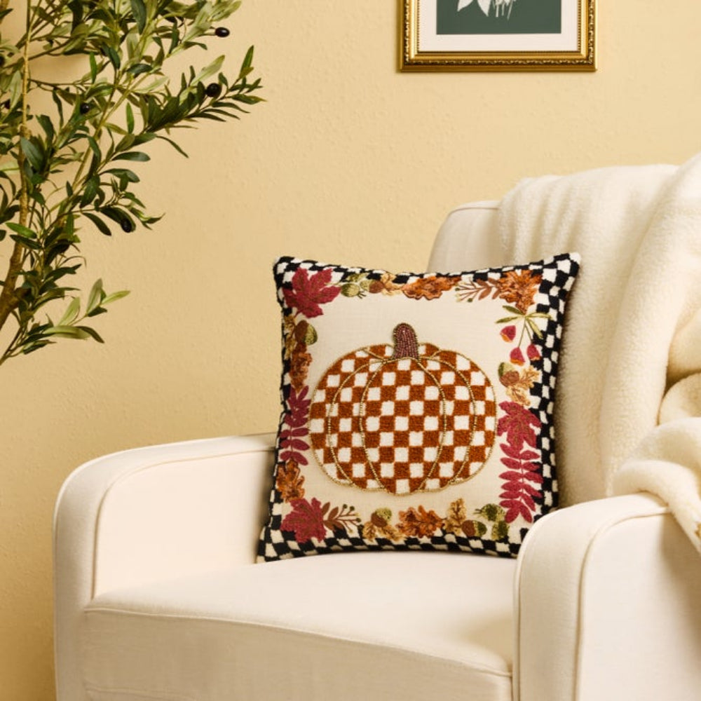 A decorative square throw pillow with a fall harvest design, resting on a cream-colored armchair in a cozy room setting.
