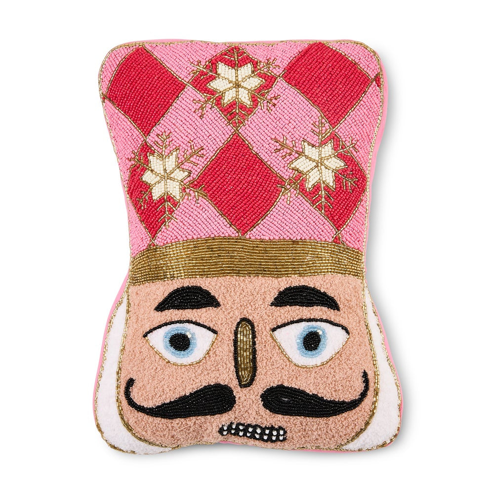 A decorative nutcracker-themed pillow featuring a pink and red diamond patterned hat with gold snowflake motifs, a gold band, and a beige face with embroidered black mustache and blue eyes.
