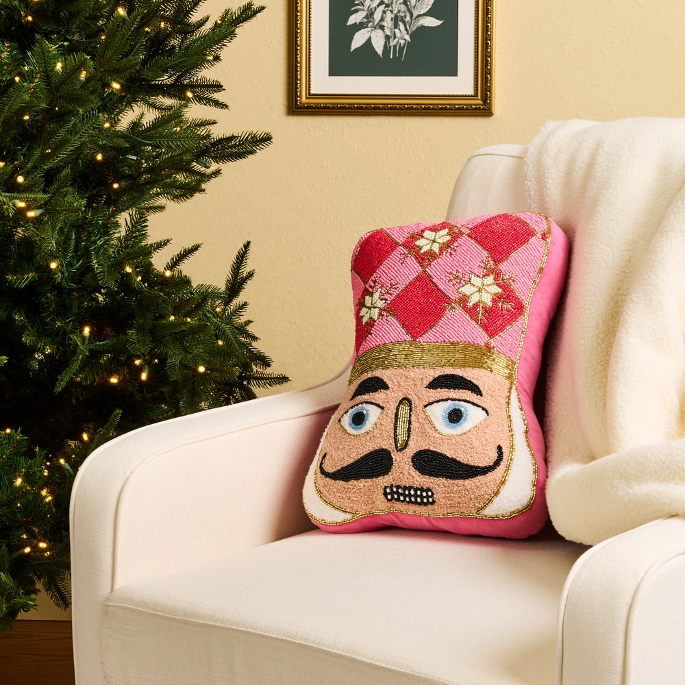 A pink nutcracker-themed throw pillow with embroidered facial features and a red-and-pink diamond patterned hat, resting on a cream armchair beside a decorated Christmas tree.
