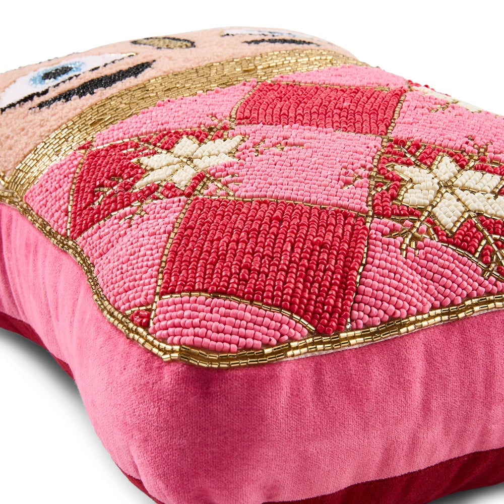 A close-up of a decorative nutcracker throw pillow featuring a pink velvet base and a chunky tufted patchwork design in shades of pink, red, and cream with gold metallic accents.