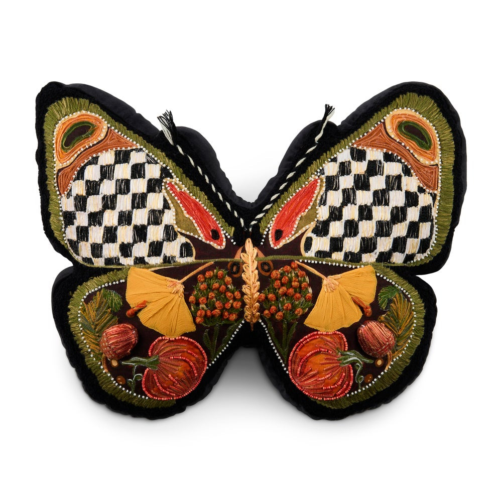 A decorative butterfly-shaped cushion or pillow with embroidered details, featuring a black and white checkerboard pattern on the upper wings, orange and red accents, and embroidered autumn motifs including pumpkins and leaves on a black background.