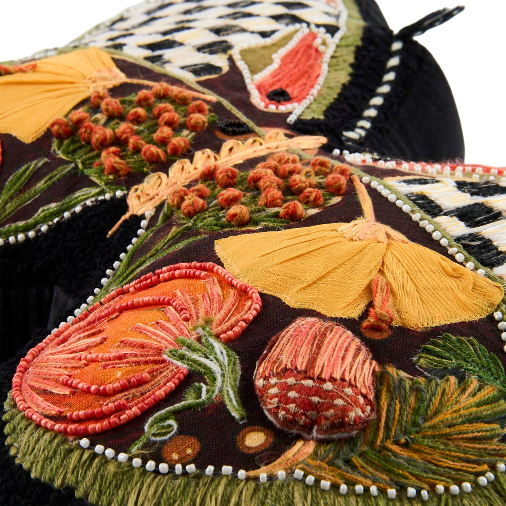 Close-up A decorative butterfly-shaped cushion or pillow with embroidered details, featuring a black and white checkerboard pattern on the upper wings, orange and red accents, and embroidered autumn motifs including pumpkins and leaves on a black background.