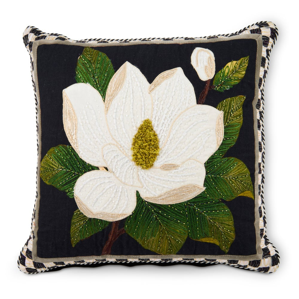 A square decorative throw pillow featuring a large white magnolia flower with green leaves on a black background, finished with a black-and-white checkered braided border.