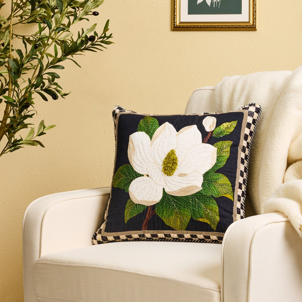 A decorative throw pillow with an embroidered white magnolia flower and green leaves on a dark navy background, styled on a cream armchair in a warm, neutral room.