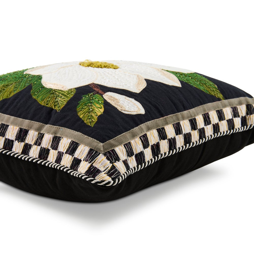 A square decorative throw pillow with a black background, featuring an embroidered white magnolia flower with green leaves on the top. The front edge has a black and white checkered border with cream fringe trim.