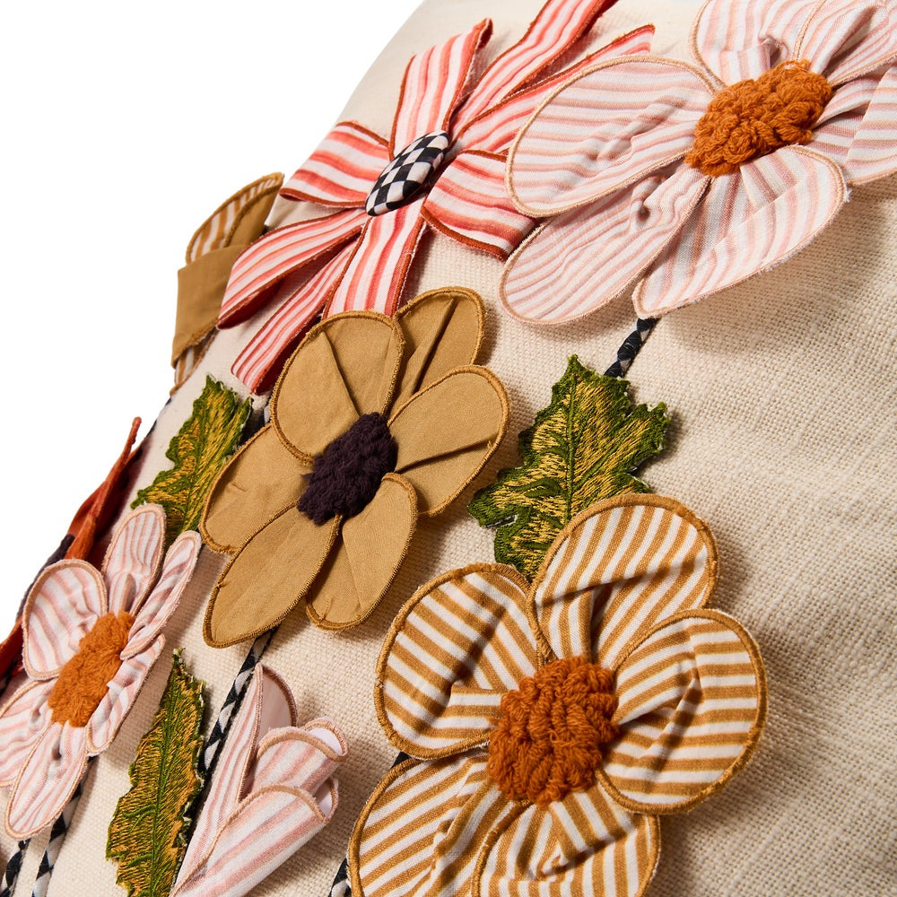 Close-up of a beige fabric surface decorated with large embroidered flowers. The flowers include striped petals in pink, cream, and orange, solid tan petals, and green leaves, with some centers in orange, dark purple, or black-and-white check.