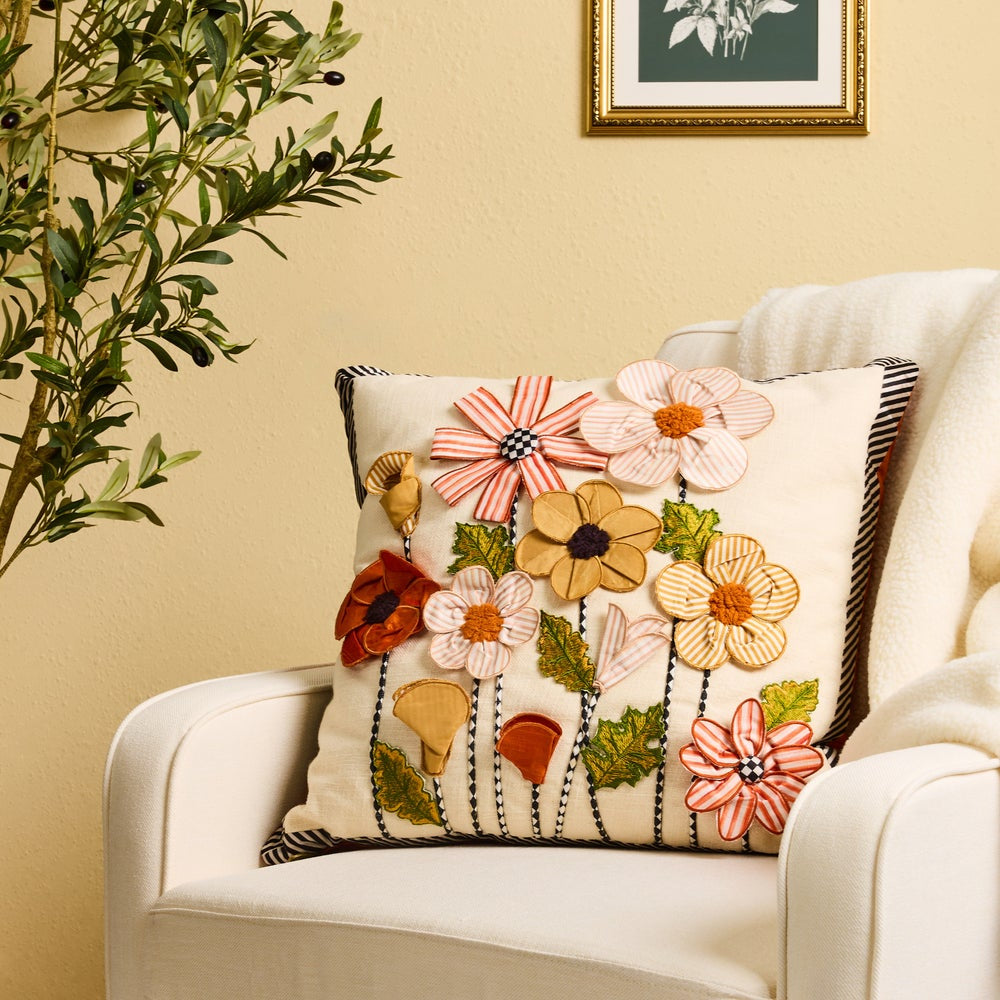 A square cream throw pillow with a 3D floral applique design, styled on a white armchair in a warm, decorated room.