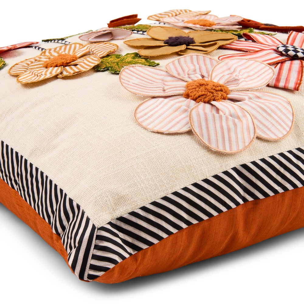 A square decorative throw pillow with a cream linen-textured center, orange fabric sides, and a black-and-white striped border. The top surface features large appliqué flowers in orange, pink-striped tones, and tan, with small green and purple fabric accents.