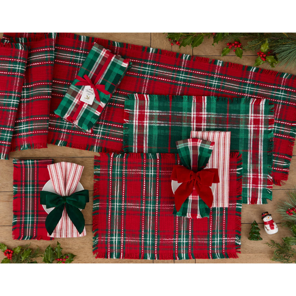 A coordinated holiday gift set arranged on a wooden surface, featuring red, green, and white plaid and striped fabrics or paper goods with fringed edges, wrapped in matching plaid and tied with bows. Christmas greenery and small decorative props are placed around the items.
