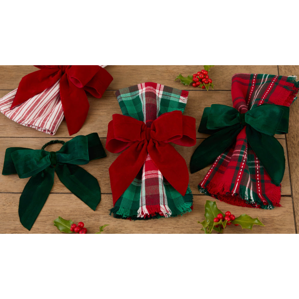 Three folded holiday napkins with large decorative bows are arranged on a wooden surface. The napkin on the left is red and white striped with a large red bow; the center napkin has a red, green, and white plaid pattern with a large red bow; the right napkin is red and green plaid with a large dark green bow.