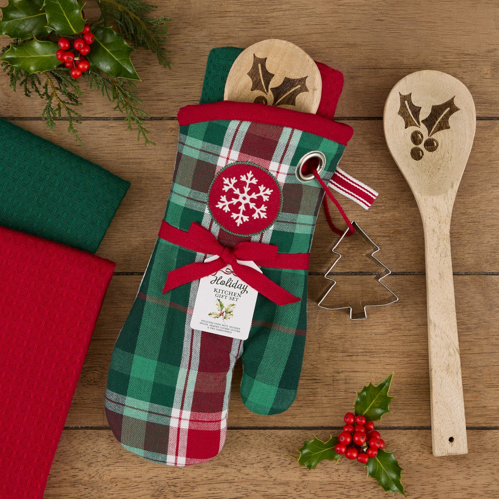 A Christmas-themed oven mitt with a green, red, and white plaid pattern, decorated with a red ribbon bow, a red snowflake patch, and a hanging tag that reads 'Holiday Kitchen Essentials.' It is styled on a wooden surface with a wooden spoon, folded red and green cloths, and holly decorations.