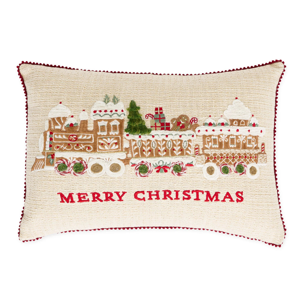 A rectangular cream-colored decorative throw pillow featuring an embroidered Christmas scene with a gingerbread train, holiday trees, candy canes, and the words 'MERRY CHRISTMAS' in red text, finished with a red pom-pom trim.