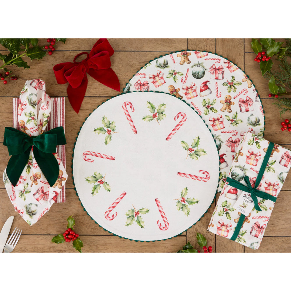 A flat-lay of a Christmas-themed tableware set on a rustic wooden table, featuring a round white platter with red and green holiday illustrations, matching napkins, and a folded napkin tied with a green ribbon.