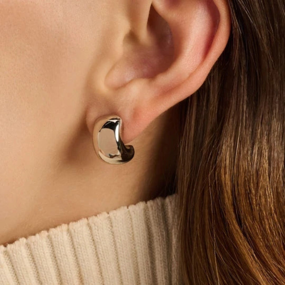 A close-up of a person's ear wearing a polished, curved gold hoop earring, paired with a cream ribbed turtleneck sweater.