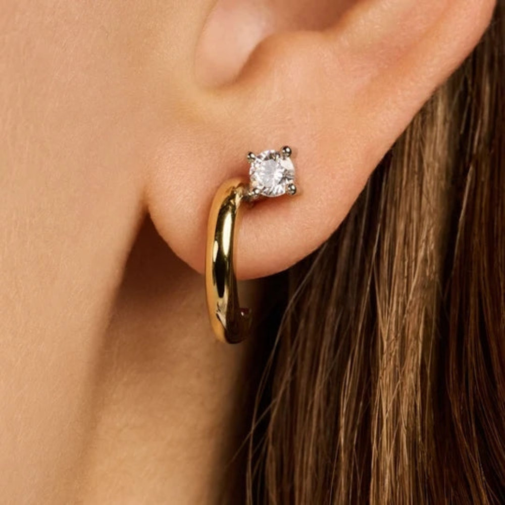 A close-up of a person's ear wearing a slim gold huggie hoop earring with a square clear gemstone in a prong setting.