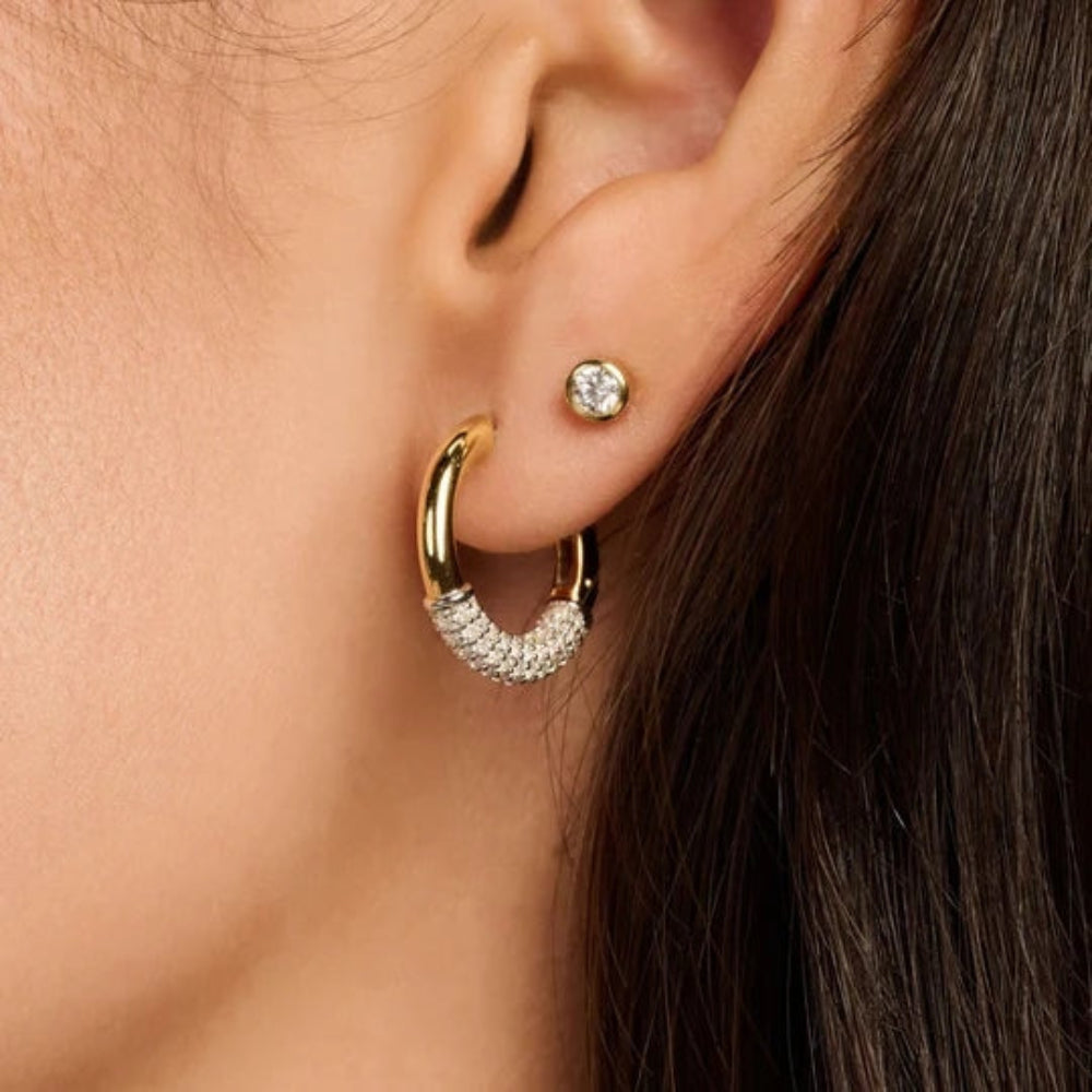A close-up of a person's ear wearing a gold hoop earring with a pavé-set curved lower section and a small round stud earring.