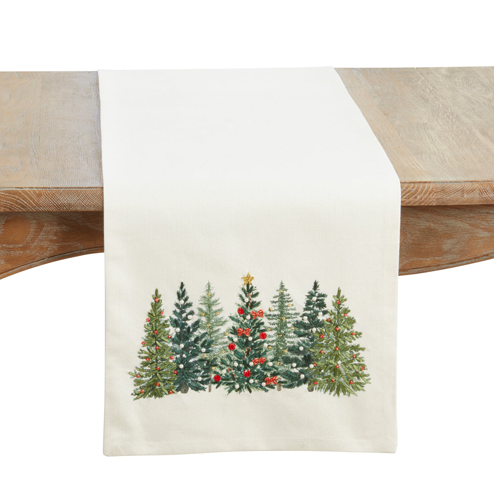 An off-white table runner featuring a printed illustration of five green Christmas trees with red berries and a gold star, draped over a rustic wooden table.
