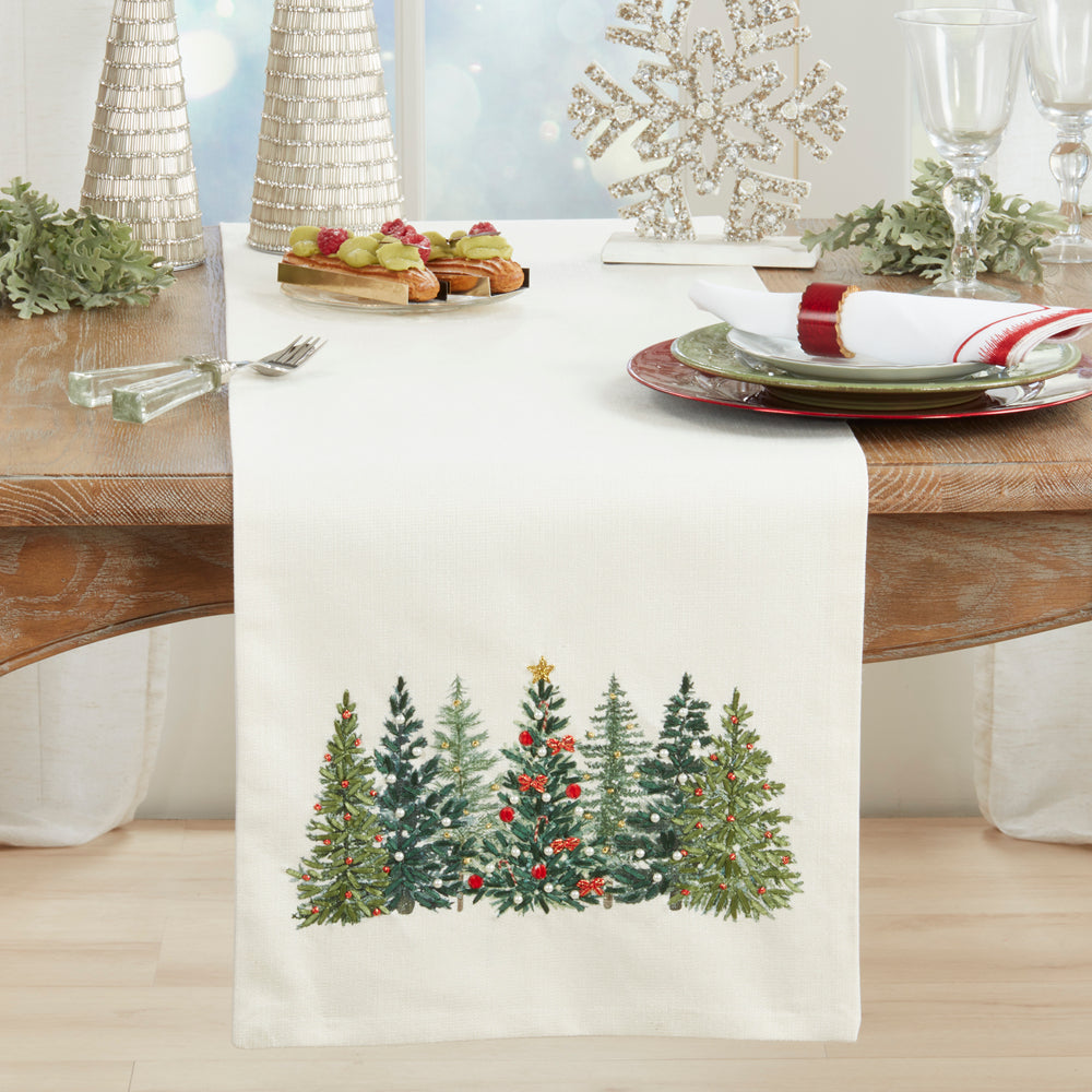 An off-white table runner with a printed row of green Christmas trees and red ornaments, styled on a wooden dining table with plates, glasses, and holiday decorations.