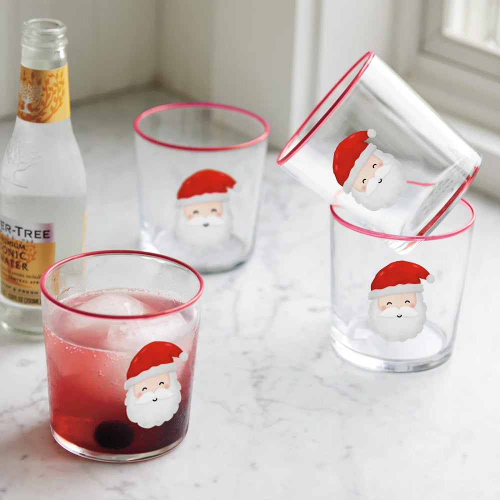 Santa themed double old fashioned glasses with santa faces on them and red rims and a glass bottle on a white marbled backround.
