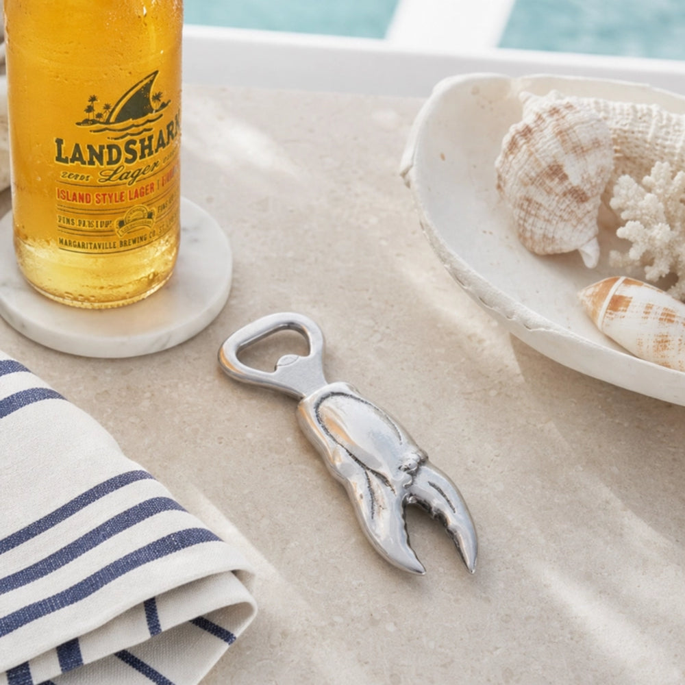 A lobster-shaped silver bottle opener resting on a light beige stone surface, styled with a Landshark beer bottle, a striped napkin, and seashells in a ceramic dish.