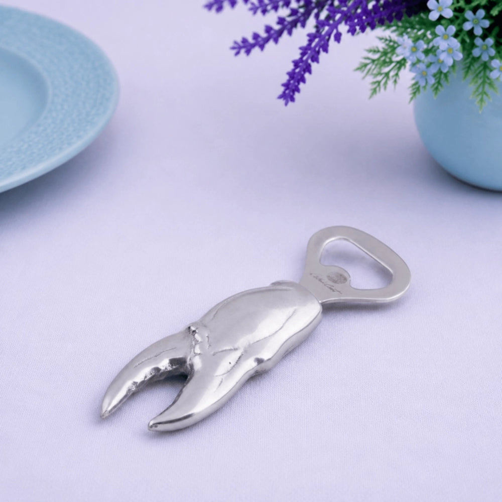 Metallic crab claw bottle opener on a light purple surface with a plate and flowers in the background.