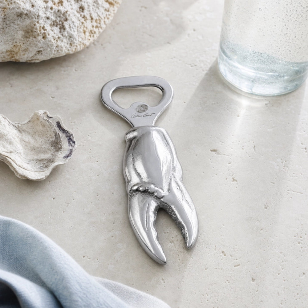 A silver metal claw-shaped bottle opener styled on a light stone surface with seashell props, a glass of water, and a blue cloth.