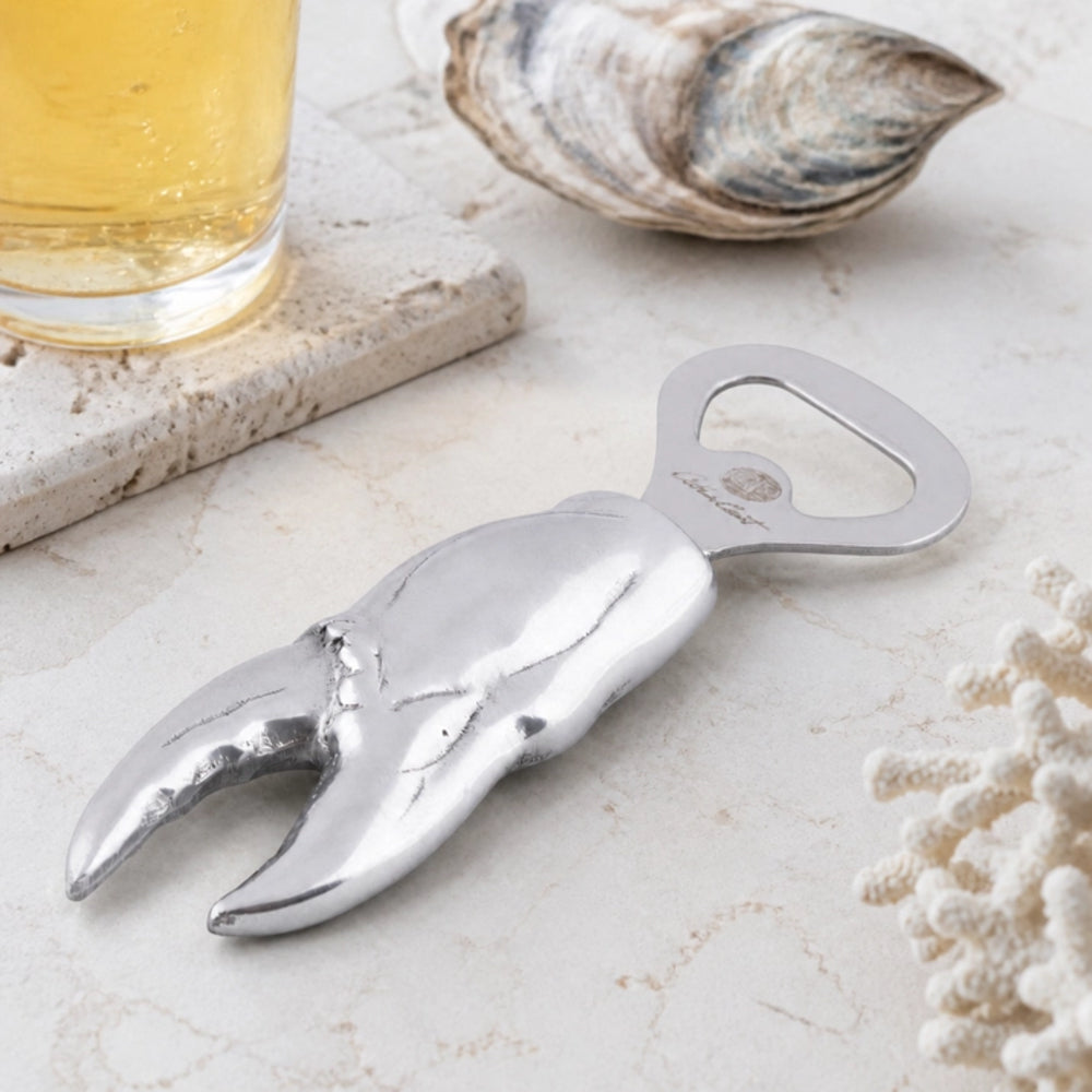 A silver lobster claw bottle opener styled on a light stone surface with a glass of beer, a clam shell, and coral as props.